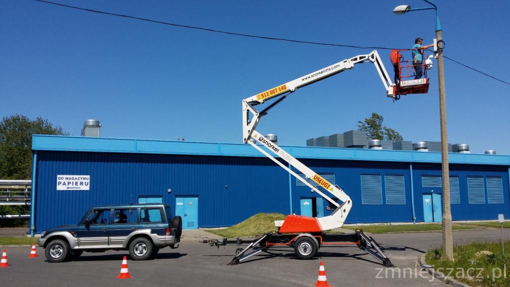 Pracownik w koszu podnośnika teleskopowego, naprawiający lampę uliczną; w tle niebieski budynek magazynu papieru i samochód terenowy.