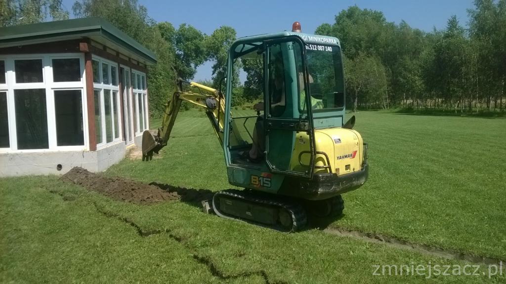 Żółto-zielona mini koparka Yanmar B15 przygotowuje wąski wykop w trawniku obok budynku z białymi oknami.