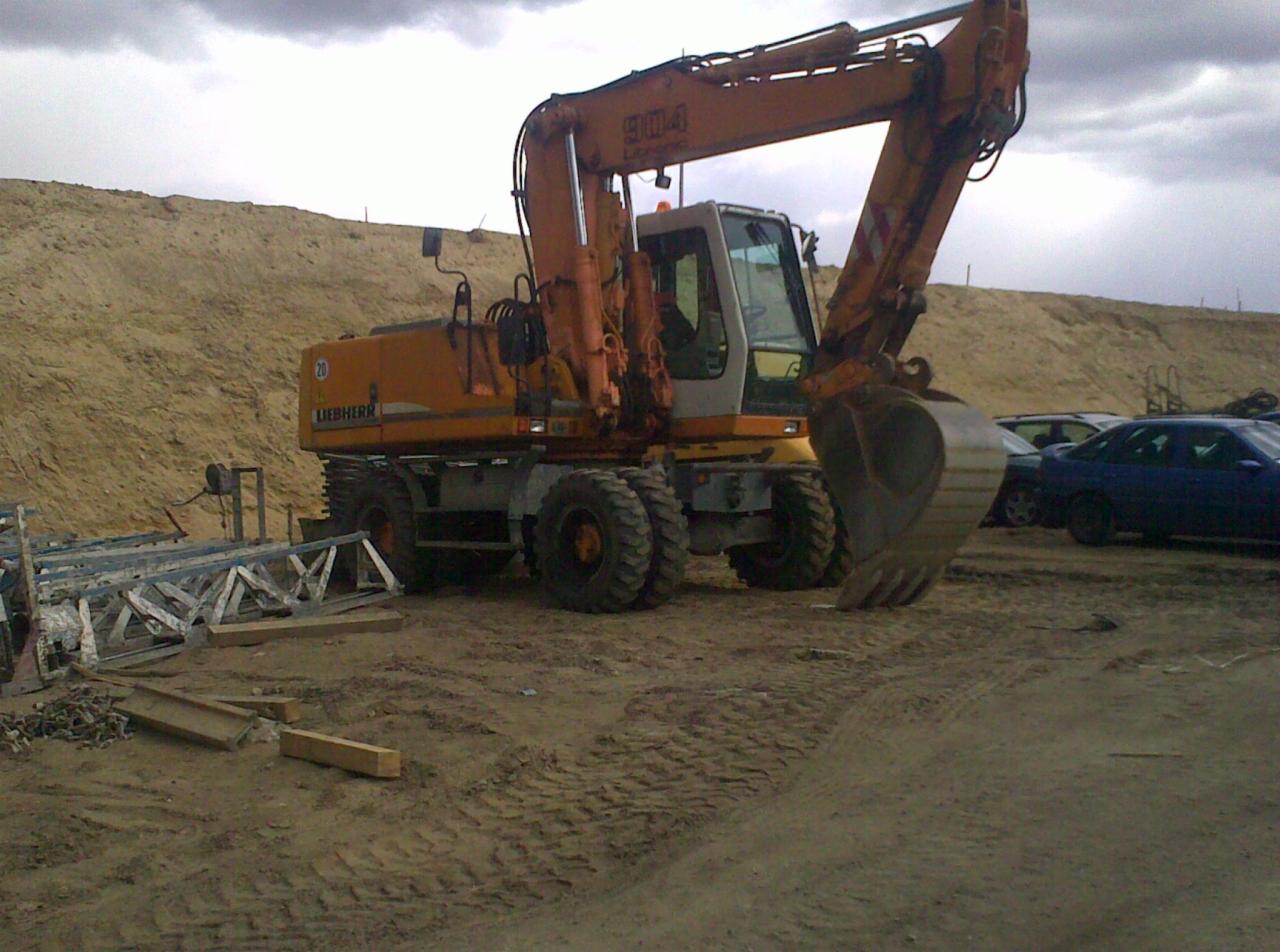 Pomarańczowa koparka Liebherr na placu budowy z fragmentami rusztowań i stertą ziemi w tle, ukazana w pochmurny dzień.