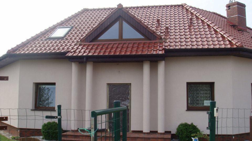 Dom jednorodzinny po remoncie dachu z czerwoną dachówką ceramiczną, widoczny zza zielonego ogrodzenia w Szczecinie.