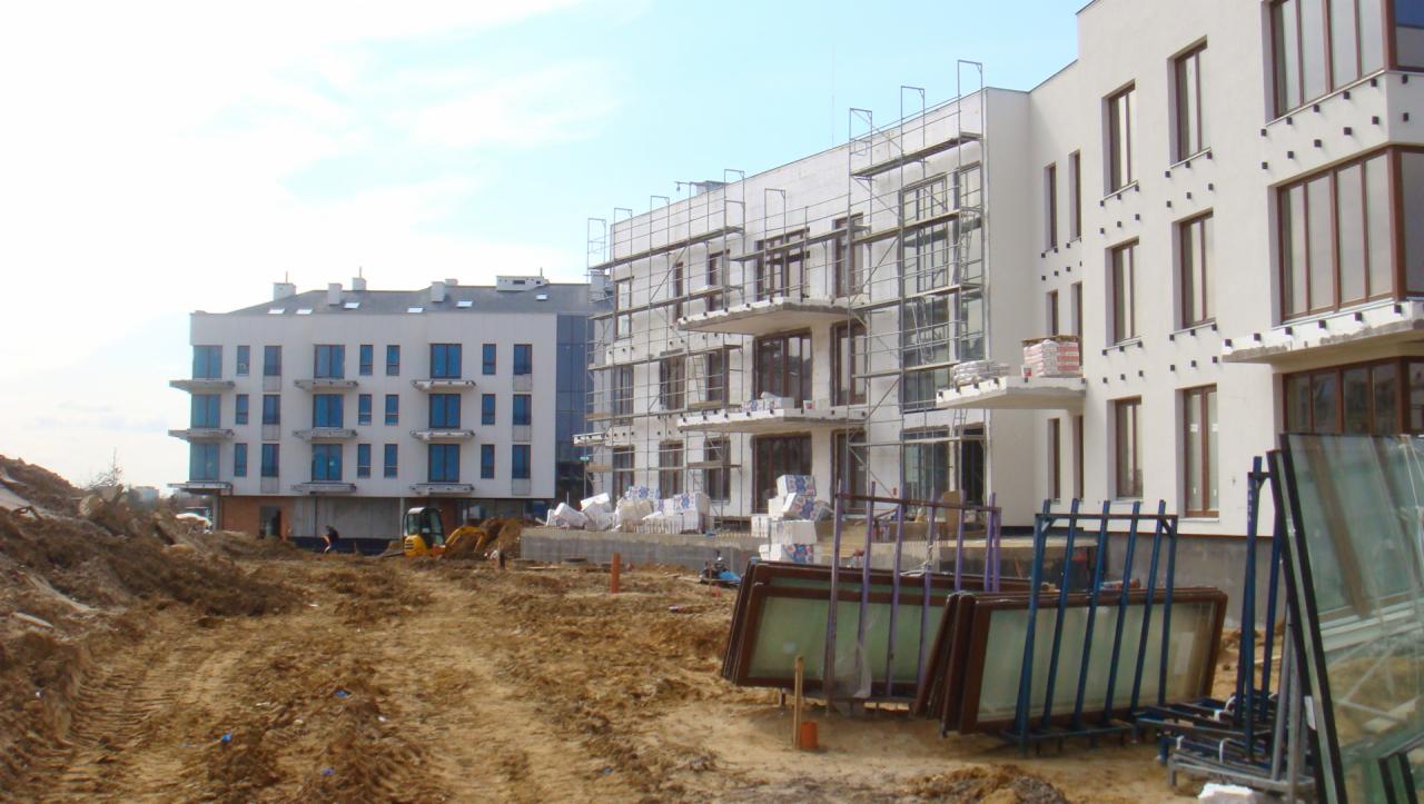 Budowa wielorodzinnego budynku z ociepleniem, widoczne rusztowania, okna czekające na montaż oraz mała koparka na placu budowy.