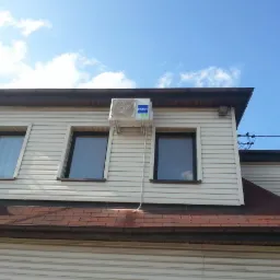 Biała jednostka zewnętrzna klimatyzacji Haier zamontowana na elewacji domu z trzema oknami, widok z dołu na tle błękitnego nieba.