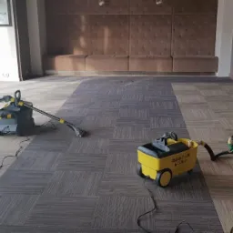 Dwa urządzenia do czyszczenia wykładzin w dużym biurze z wykładziną w szaro-beżową kratę; część wykładziny jest już wyczyszczona i ma ciemniejszy odcień.