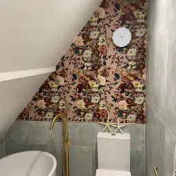 Ukończona łazienka na poddaszu z kwiatową tapetą na skośnej ścianie, szare płytki imitujące marmur, biała wanna wolnostojąca i złota bateria podłogowa, dwie białe rozgwiazdy na spłuczce toalety.