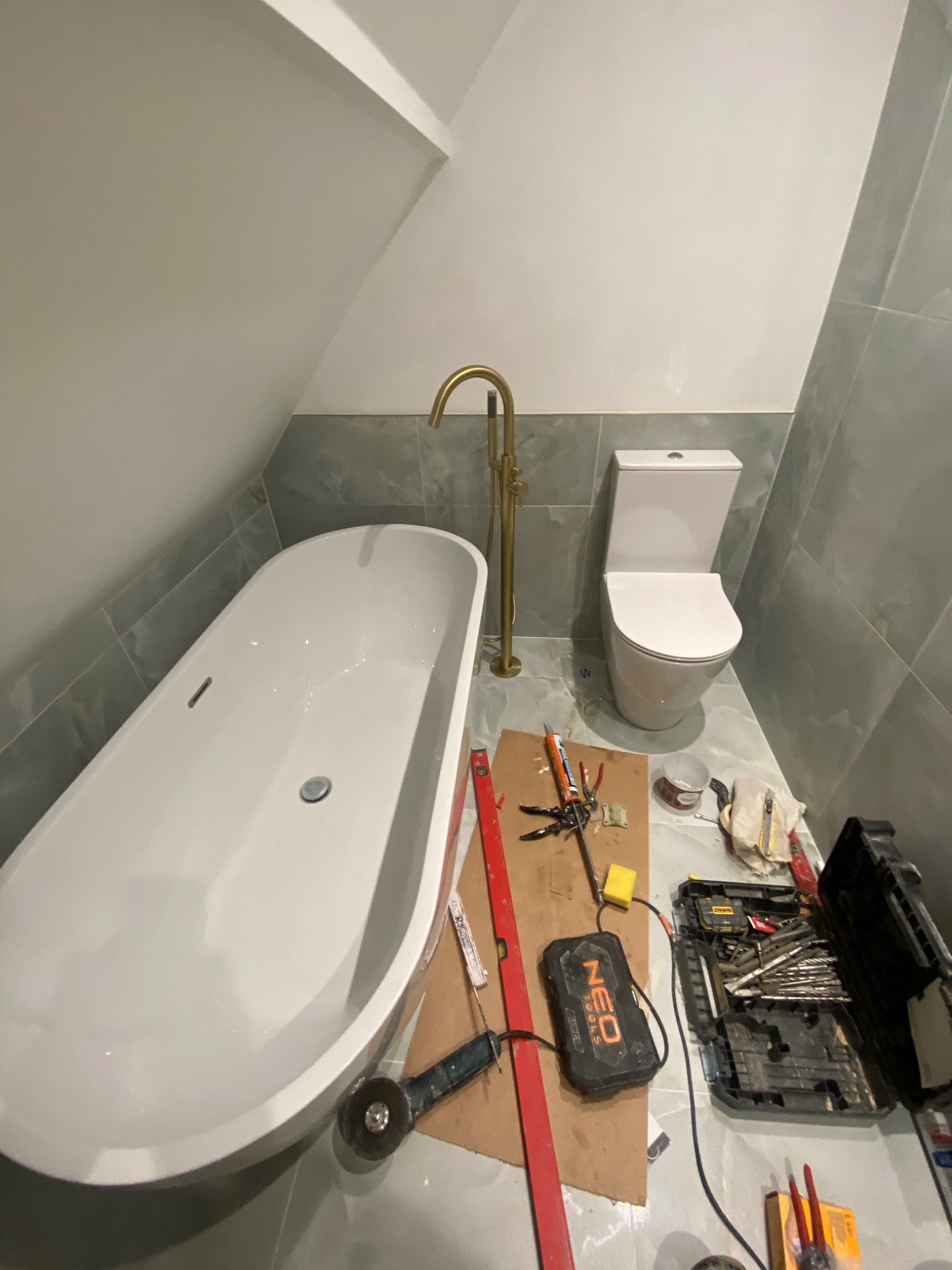 Montaż armatury w łazience na poddaszu: wolnostojąca wanna z baterią w kolorze złotym, toaleta i narzędzia glazurnicze na podłodze wyłożonej szarymi płytkami.