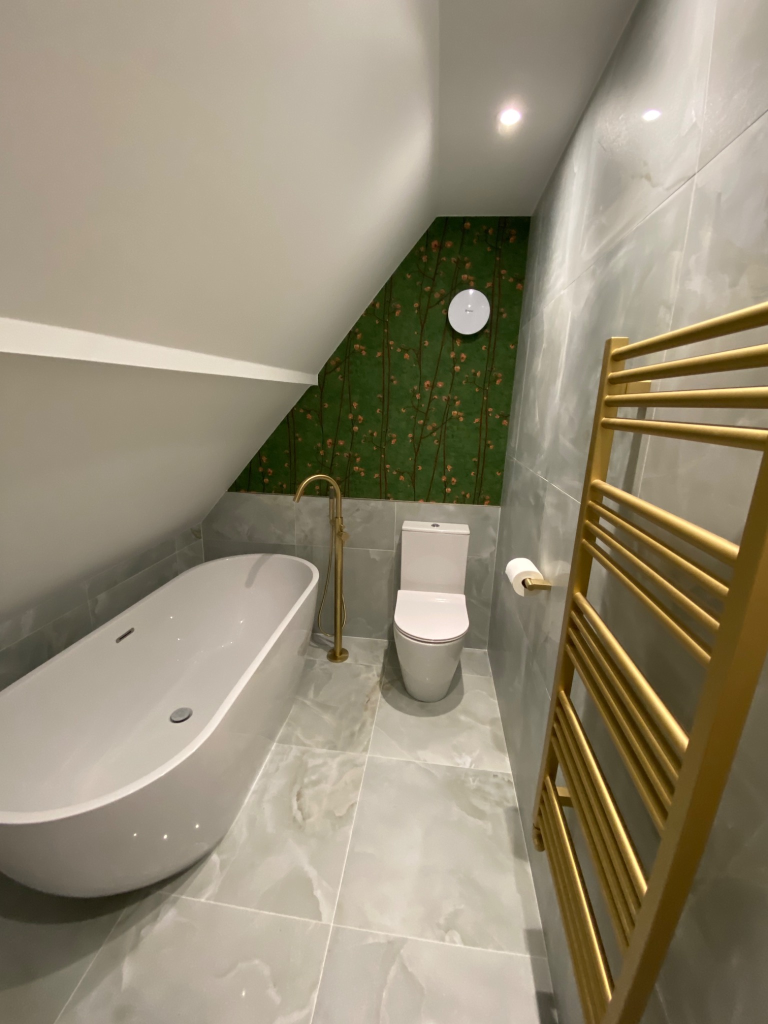 Nowoczesna łazienka na poddaszu z wanną wolnostojącą, złotą armaturą, grzejnikiem drabinkowym w kolorze złota, toaletą i zieloną tapetą w kwiaty na skośnej ścianie