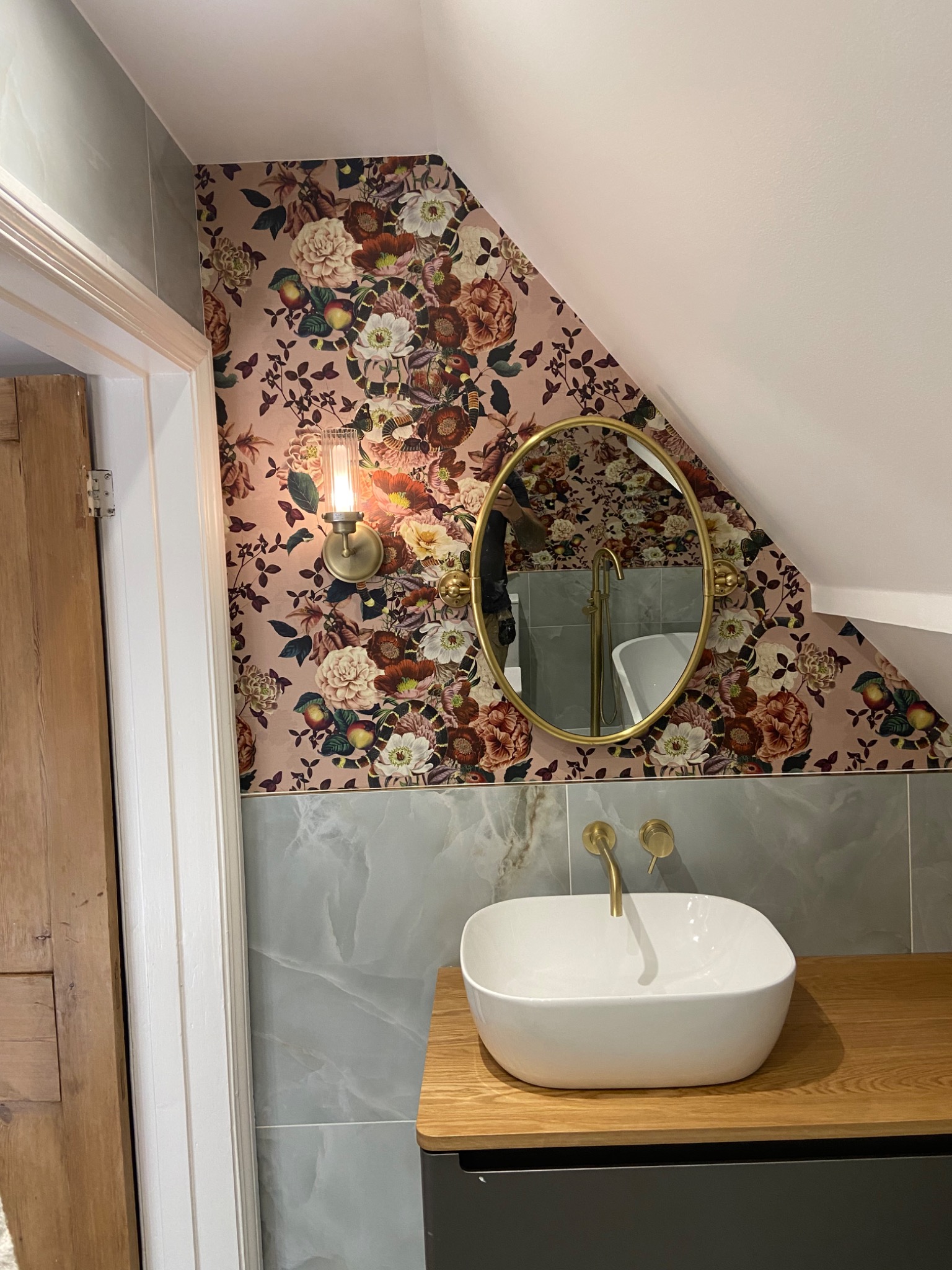 Ukończona łazienka w stylu retro z umywalką nablatową, złotą armaturą i owalnym lustrem w złotej ramie na tle ściany z tapetą w kwiaty i szarymi płytkami imitującymi marmur.