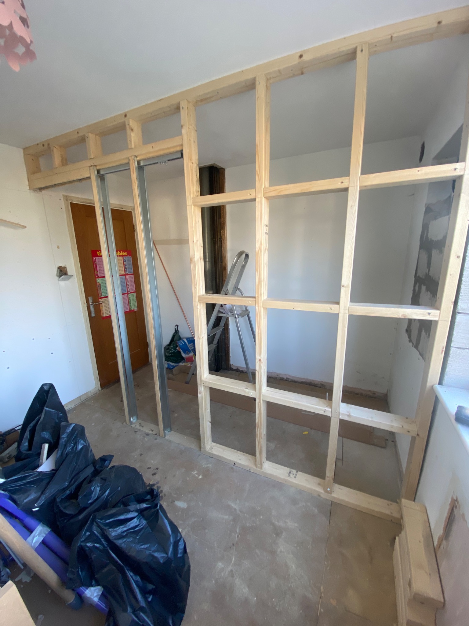 Konstrukcja szkieletowa z drewna i metalu w trakcie budowy ścianki działowej w pomieszczeniu, widoczne materiały budowlane i drabina.