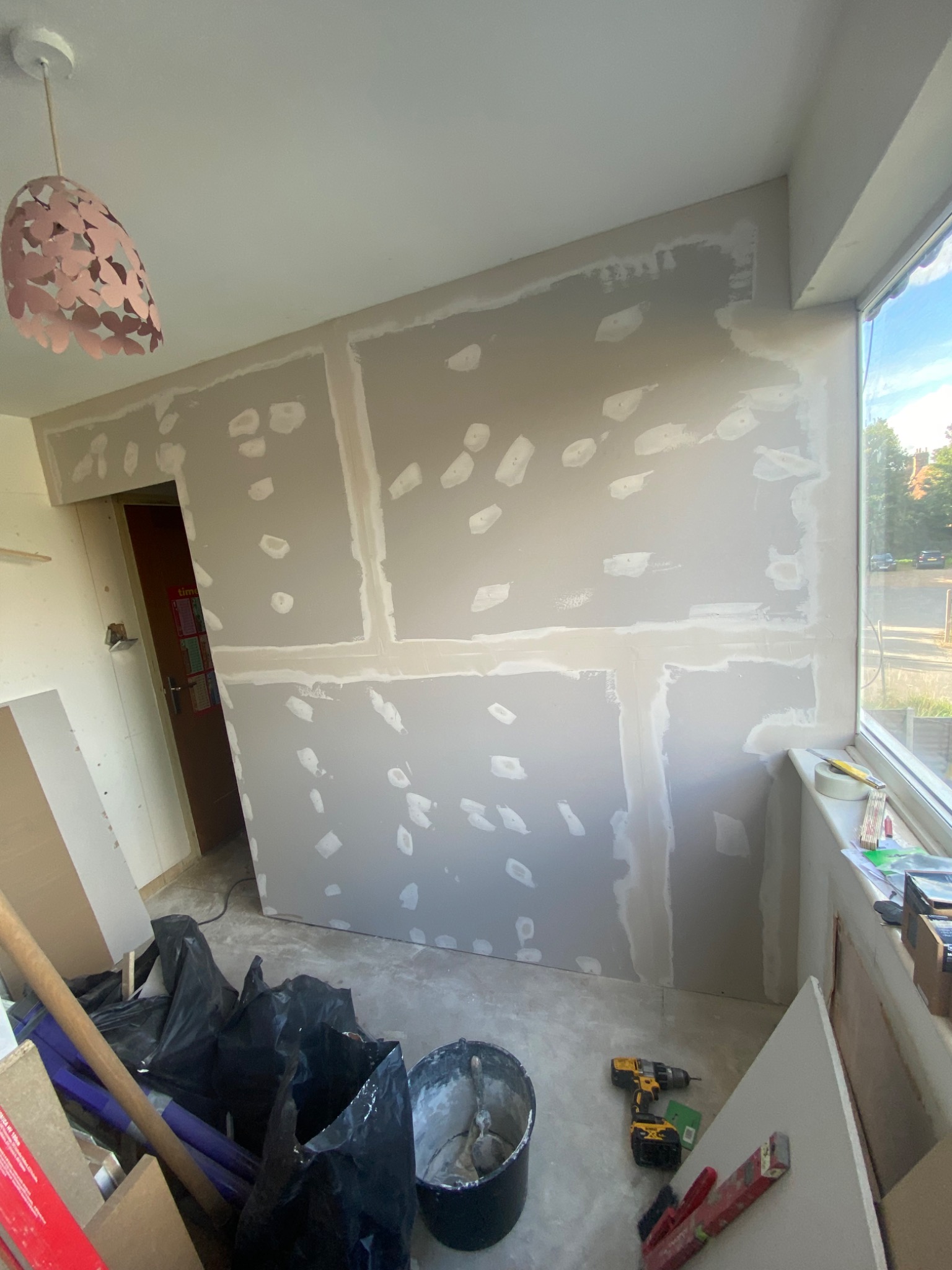 Pokój w trakcie remontu: ściany z płyt gipsowo-kartonowych z widocznymi łączeniami i miejscami po szpachlowaniu, narzędzia budowlane w nieładzie na podłodze, plastikowe worki, w tle widoczne okno...