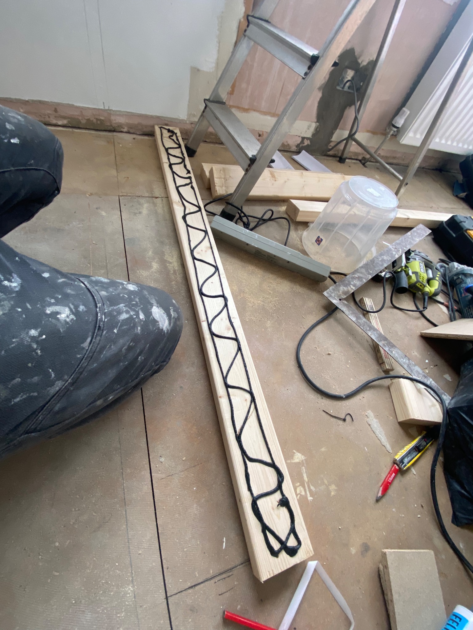 Deska z zygzakowatą warstwą czarnego kleju leżąca na podłodze, obok ubrudzonych farbą spodni roboczych, drabiny i narzędzi w trakcie prac remontowych.