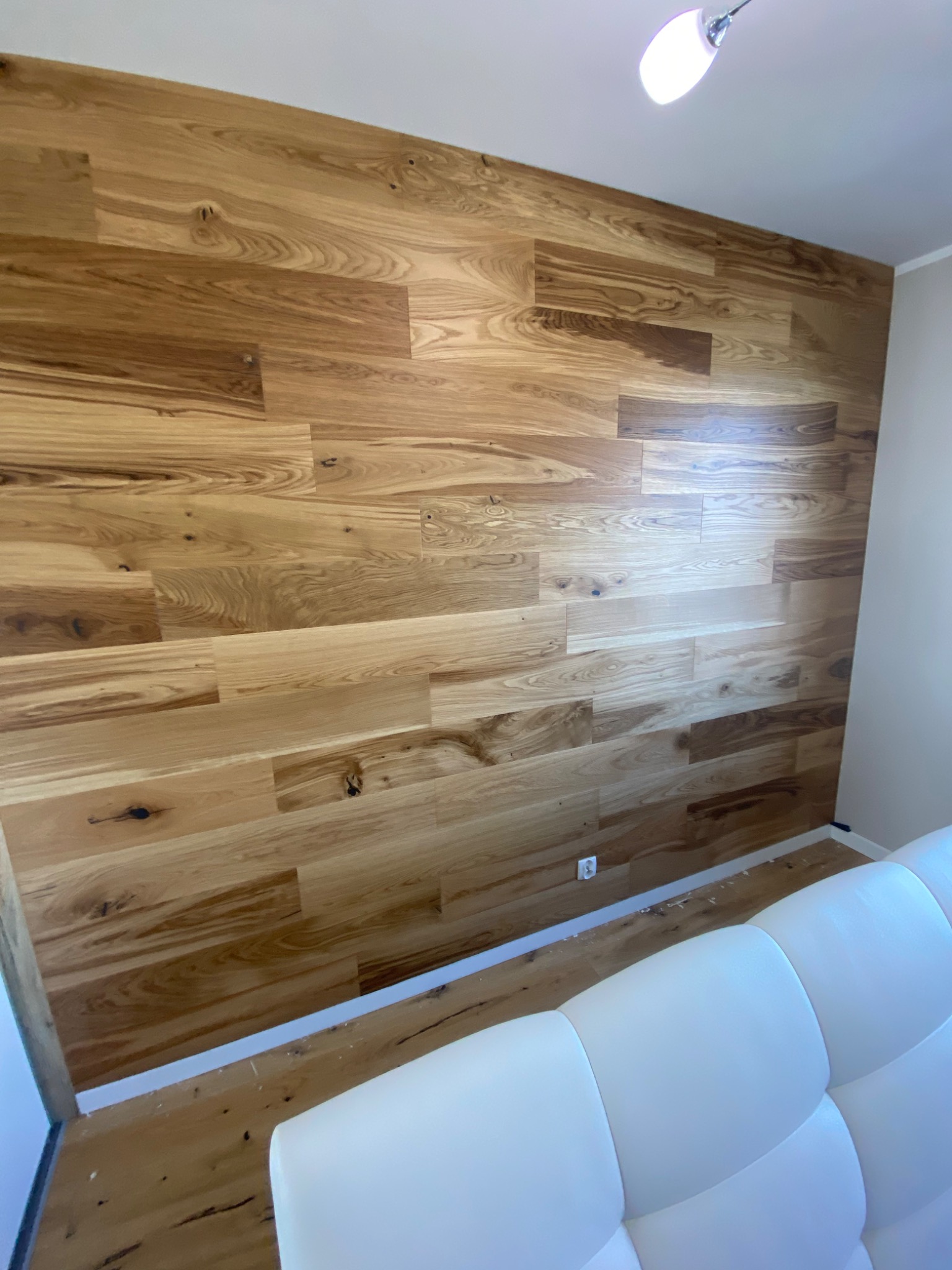 Ściana wyłożona panelami z naturalnego drewna o ciepłym odcieniu, widoczne słoje i sęki, fragment białej sofy w dolnym rogu, oświetlenie sufitowe.