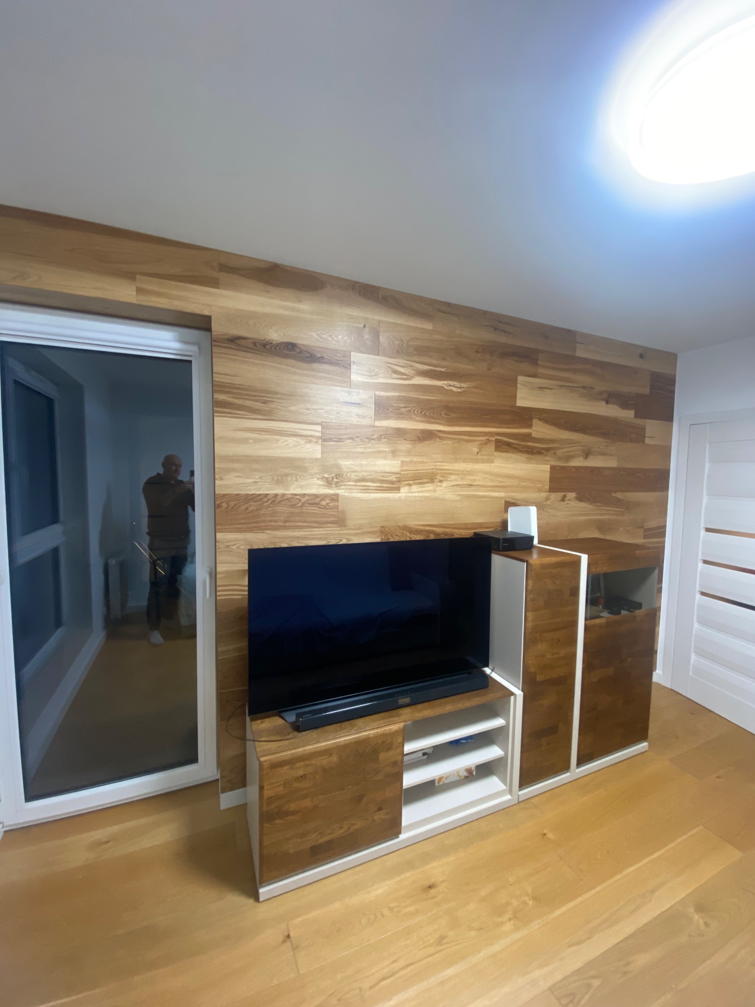 Drewniana ściana z telewizorem i szafką RTV, widok na balkon przez przesuwne drzwi, podłoga z paneli drewnianych, odbicie fotografa w szybie.