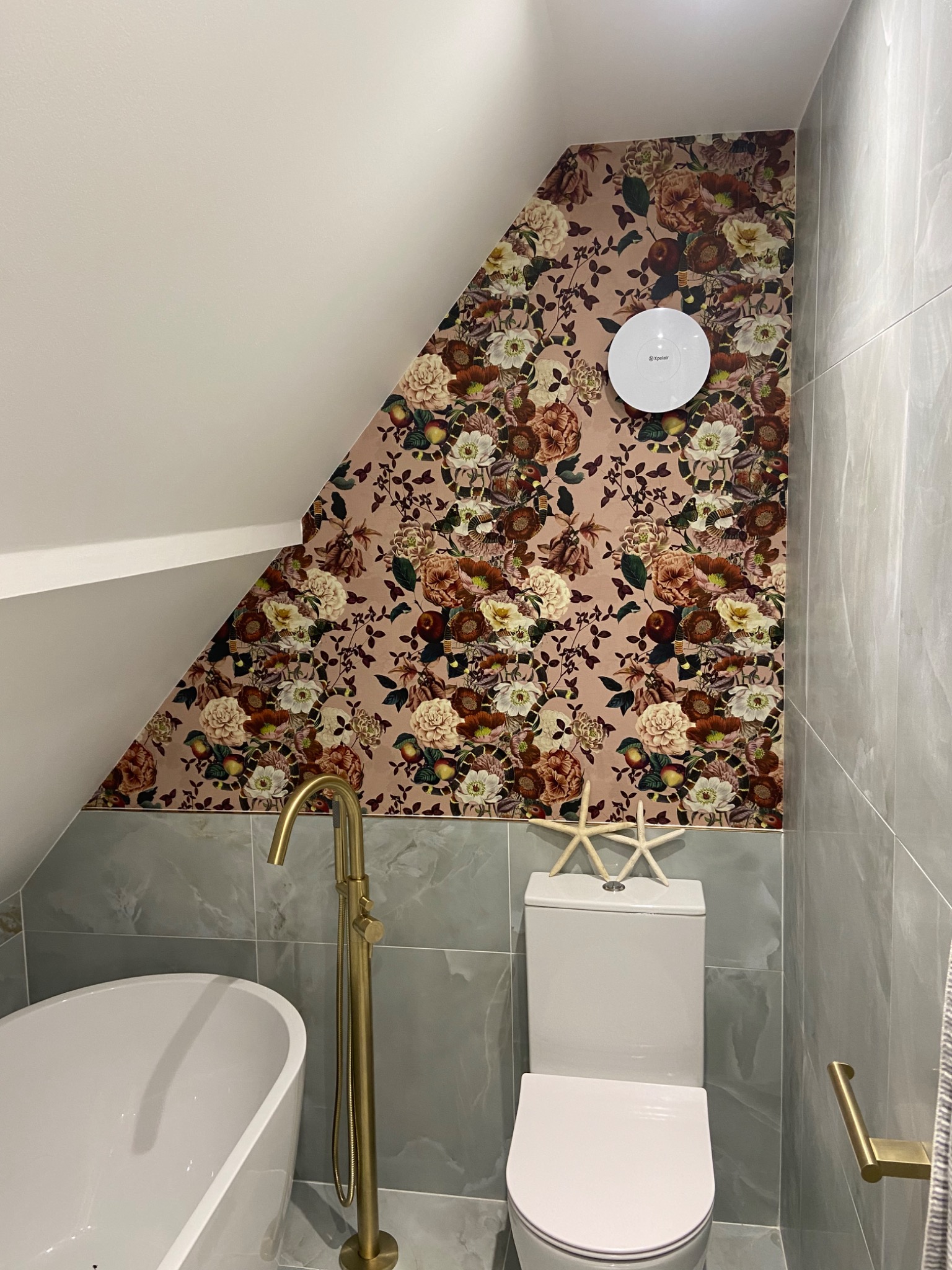 Ukończona łazienka na poddaszu z kwiatową tapetą na skośnej ścianie, szare płytki imitujące marmur, biała wanna wolnostojąca i złota bateria podłogowa, dwie białe rozgwiazdy na spłuczce toalety.