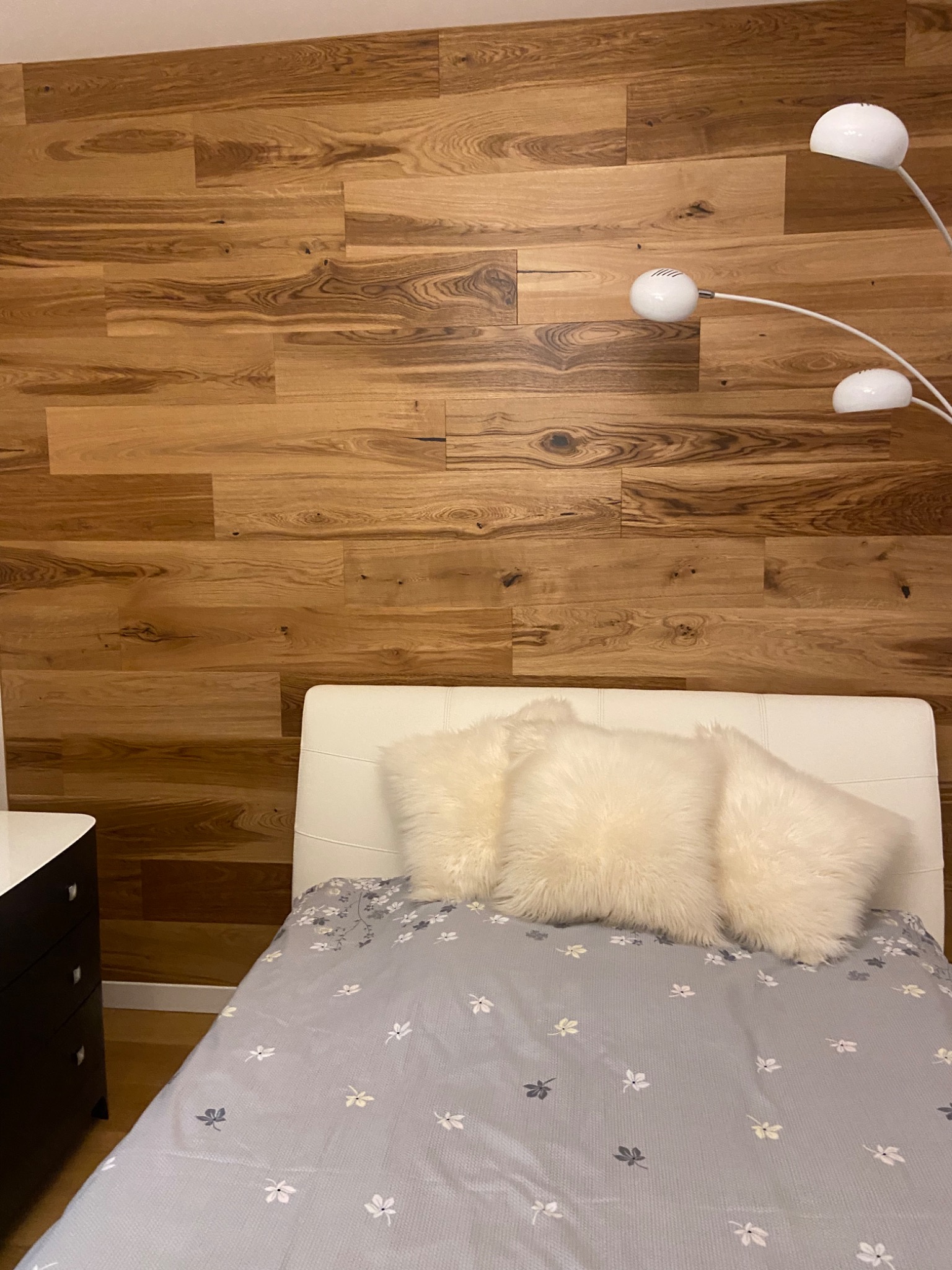 Drewniana ściana z paneli za wezgłowiem łóżka z białymi poduszkami i lampą podłogową z trzema kulistymi kloszami.