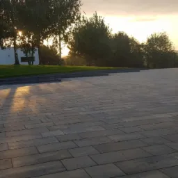 Rozległy plac wyłożony prostokątną, szarą kostką brukową, zarys budynku z białymi ścianami i zielonym trawnikiem w tle, oświetlony promieniami zachodzącego słońca, z widoczną przyczepą po prawej...