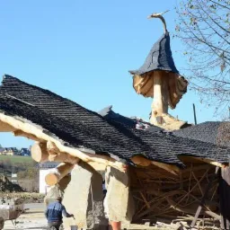 Nietypowy dach z drewnianą konstrukcją w kształcie grzyba, pokryty czarnym gontem bitumicznym, w tle krajobraz z domami i pracownik wykonujący prace wykończeniowe.