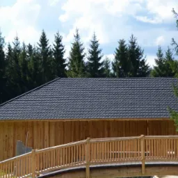 Ciemnoszara dachówka bitumiczna na drewnianym budynku z bali, widok z drewnianego mostku na tle lasu iglastego i błękitnego nieba z chmurami.