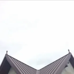 Szczyt dachu pokryty blachą trapezową w kolorze brązowym, zwieńczony dekoracyjnym elementem, na tle pochmurnego nieba.