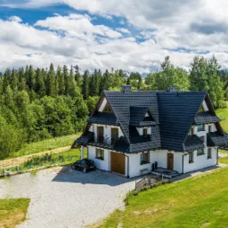 Dom jednorodzinny z ciemną dachówką, białymi ścianami i balkonami, otoczony zielonym trawnikiem i lasem, widok z lotu ptaka.