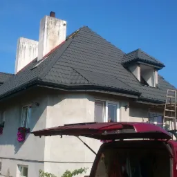 Dom z ciemnoszarym dachem z blachy trapezowej. Widoczne okna dachowe, rynny i komin. Na rusztowaniu wiadro. Częściowo widoczny samochód ekipy dekarskiej.