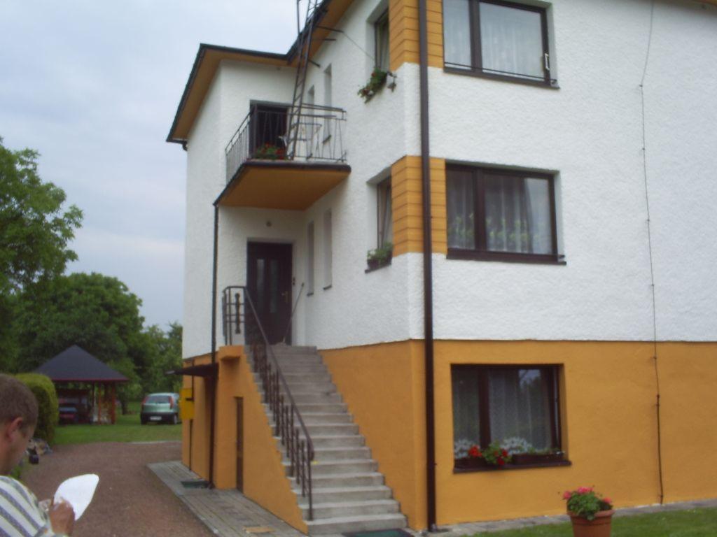 Dwukondygnacyjny budynek z elewacją w kolorach białym i żółtym, schody prowadzące do wejścia na piętrze, balkon z czarną balustradą, widoczny fragment osoby trzymającej dokumenty w lewym dolnym rogu.