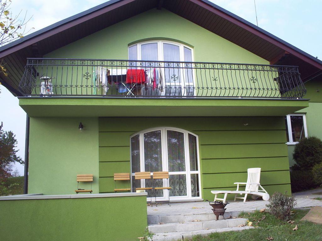 Dwupiętrowy dom z elewacją w odcieniach zieleni, z balkonem z czarną, ozdobną balustradą, na którym suszą się ubrania. Na parterze białe, łukowe okno i kilka drewnianych krzeseł oraz biały leżak.