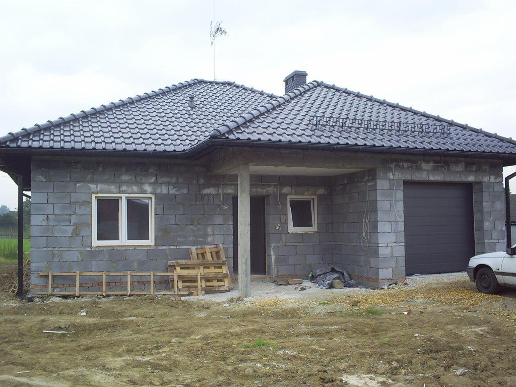 Surowy stan domu jednorodzinnego z szarej kostki betonowej, czarny dach z dachówki ceramicznej, widoczne palety budowlane, teren wokół w trakcie przygotowania.