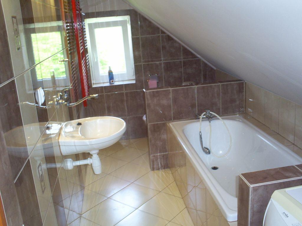 Ukończona łazienka na poddaszu z brązowymi płytkami, białą wanną i umywalką, widok na okno zza grzejnika.