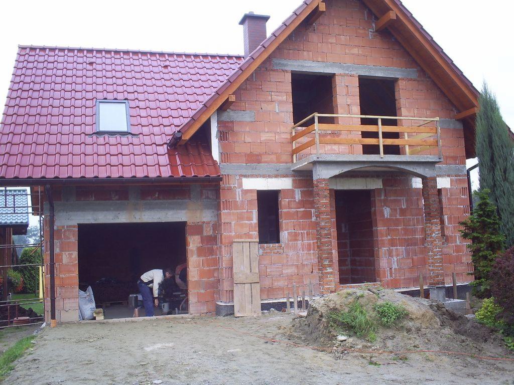 Budowa domu jednorodzinnego w trakcie realizacji: widoczna konstrukcja z czerwonej cegły, dach z czerwonej dachówki, niezadaszony balkon z drewnianą barierką, pracownik wewnątrz garażu.