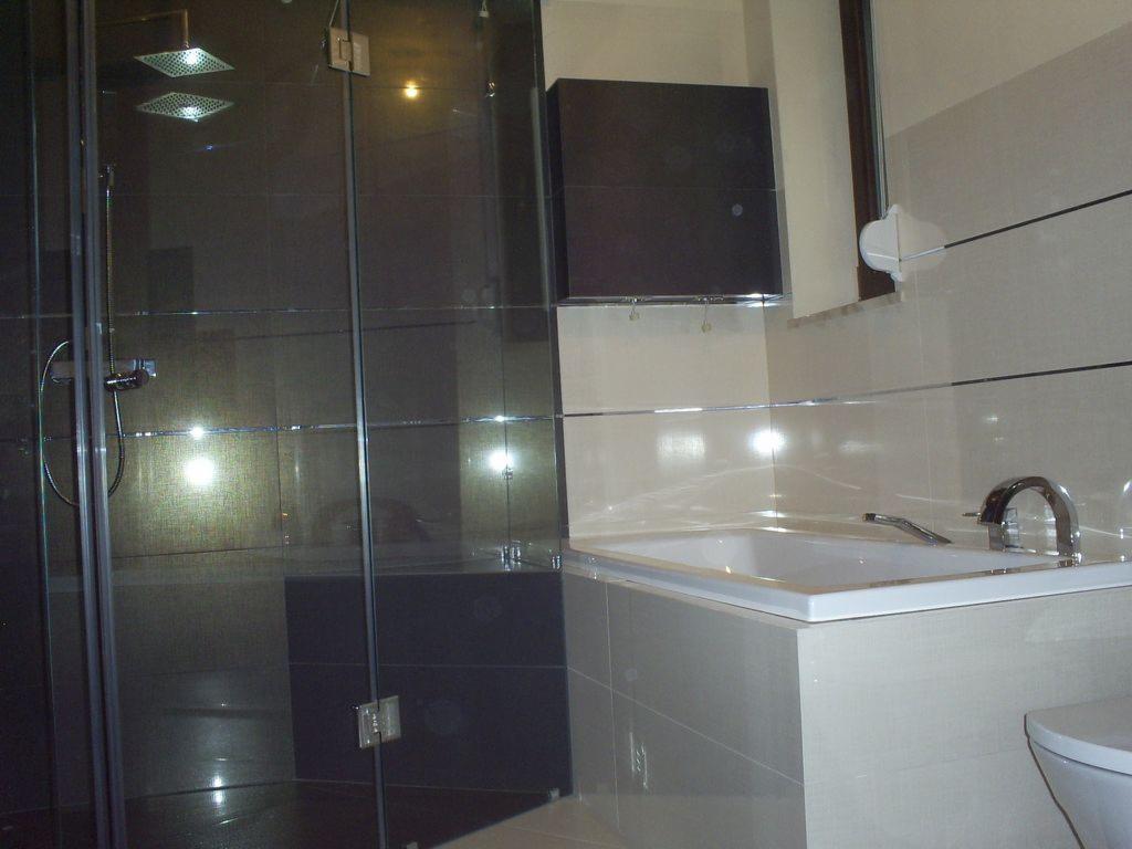 Nowoczesna łazienka z kabiną prysznicową typu walk-in z ciemnymi płytkami, białą wanną obudowaną jasnymi płytkami i chromowaną armaturą.