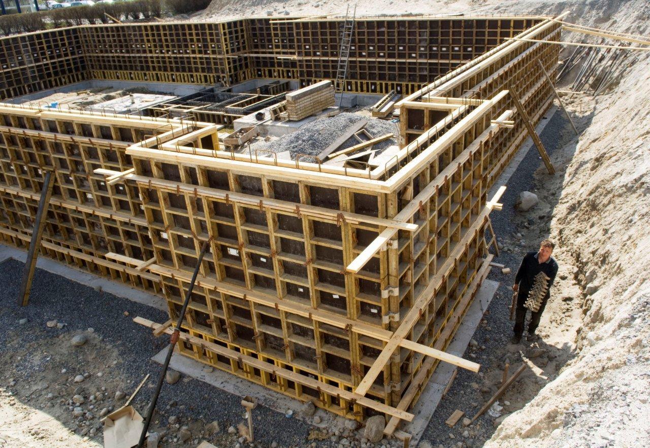 Szalunek systemowy z drewnianą konstrukcją kratową, przygotowany do wylania fundamentów, widoczny wykop i pracownik trzymający element szalunku.