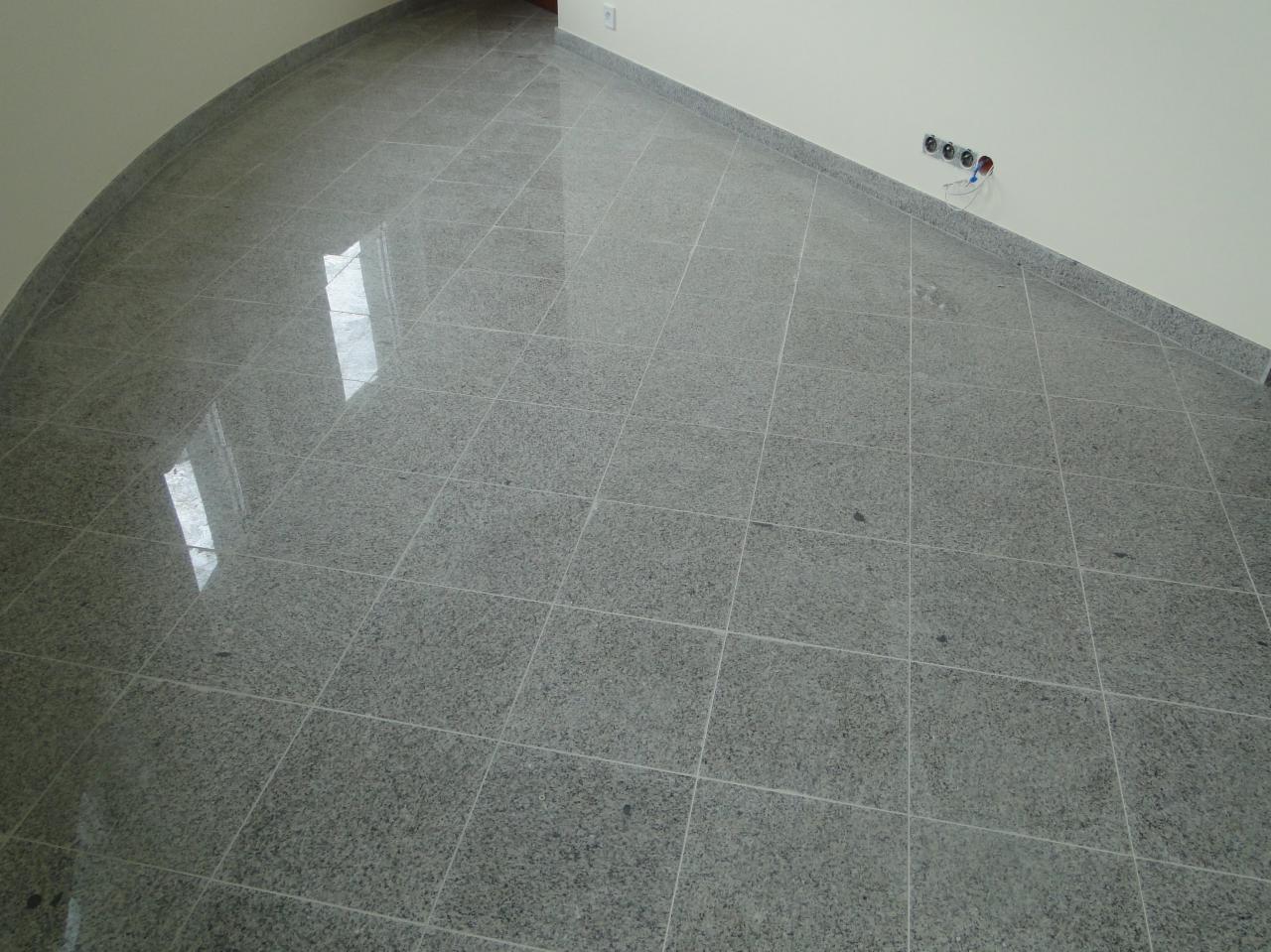 Szara, błyszcząca podłoga z płytek w kształcie kwadratów, ułożona w pomieszczeniu o zaokrąglonym narożniku. Widoczne gniazdka elektryczne przy ścianie.