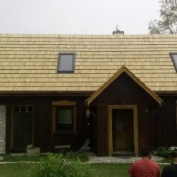 Drewniany dom z nowym dachem krytym gontem drewnianym, z oknami dachowymi. Dekoracyjne wykończenia okien i wejścia. Część budynku z kamienia, reszta z ciemnego drewna.