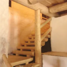 Wnętrze domu z bali: schody drewniane z balustradą z surowego drewna, okrągłe belki stropowe i ściany w jasnych odcieniach tworzą rustykalny klimat.