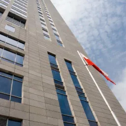 Elewacja biurowca z flagą Polski, widok z dołu. Szare płytki elewacyjne, regularny układ okien. Błękitne niebo z chmurami w tle. Ujęcie pod kątem.