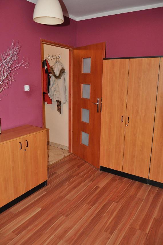 Wnętrze mieszkania z drewnianą podłogą, szafą, komodą i drzwiami z szybkami, ściana w kolorze fuksji, widok na przedpokój z wieszakami.