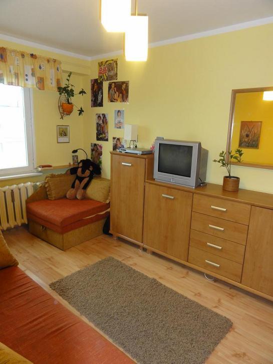 Jasne wnętrze pokoju z żółtymi ścianami, drewnianą podłogą i meblami, dywanem oraz telewizorem na komodzie. Na ścianie wiszą plakaty i lustro.