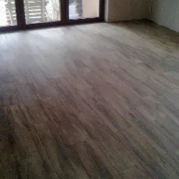 Ułożona podłoga z paneli imitujących drewno w nowym wnętrzu. Widoczne okno balkonowe i fragment ściany. Układanie paneli podłogowych.