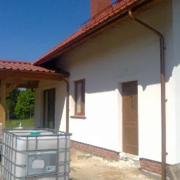 Boczny widok otynkowanego domu z brązową stolarką okienną i drewnianą wiatą. Na pierwszym planie zbiornik na wodę i przyczepka.