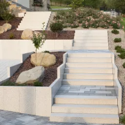 Zewnętrzne schody z jasnych płyt betonowych prowadzące przez ozdobne rabaty z roślinami i kamieniami, widok na ogród.