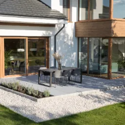 Nowoczesny taras z szarych płyt, białego żwiru i zielonego trawnika przylegający do domu z dużymi oknami.