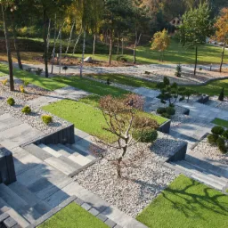 Rozległy, wielopoziomowy ogród z szarej kostki, trawników i kamiennych rabat z drzewami, widziany z góry w słoneczny dzień.