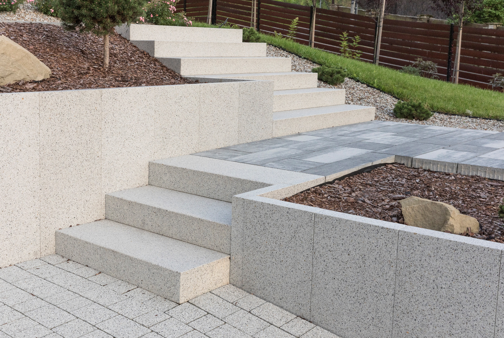 Schody zewnętrzne z jasnych, szarych płyt z drobnym wzorem, prowadzące na wyższy poziom ogrodu, obok mur oporowy i nawierzchnia z kostki w odcieniach szarości.