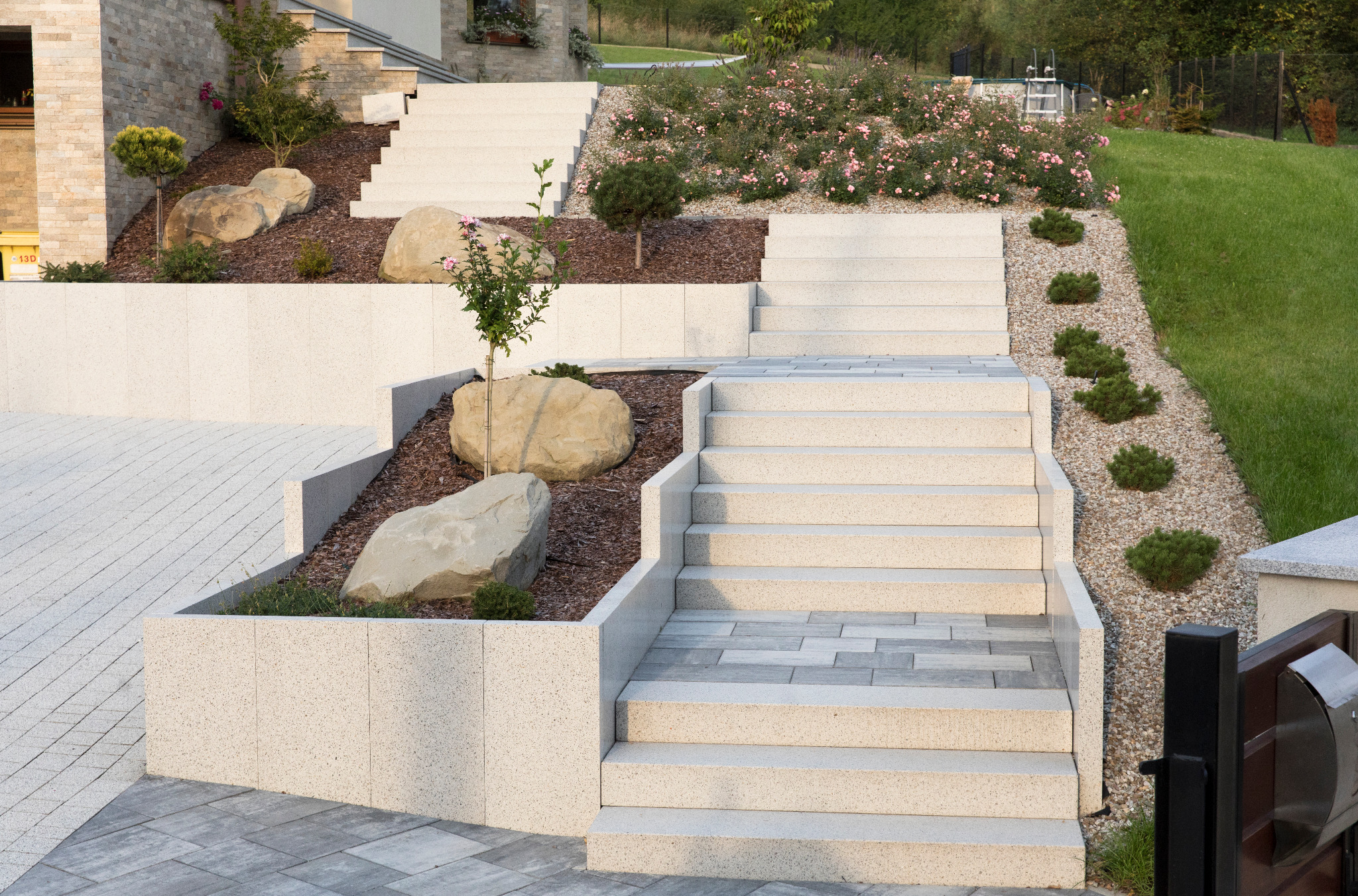 Zewnętrzne schody z jasnych płyt betonowych prowadzące przez ozdobne rabaty z roślinami i kamieniami, widok na ogród.