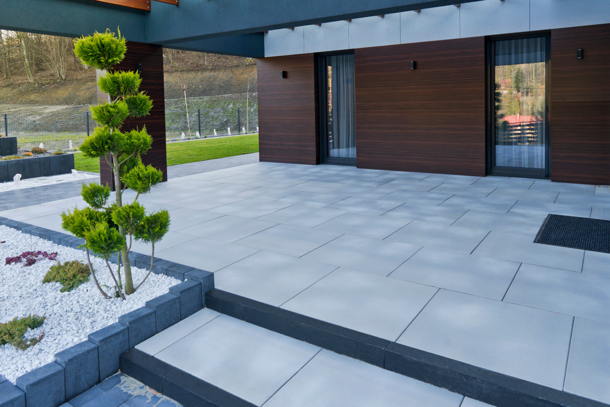 Nowoczesny taras z szarych płyt, obrzeża z ciemnego kamienia, ozdobne drzewko iglaste i widok na zadbany trawnik.