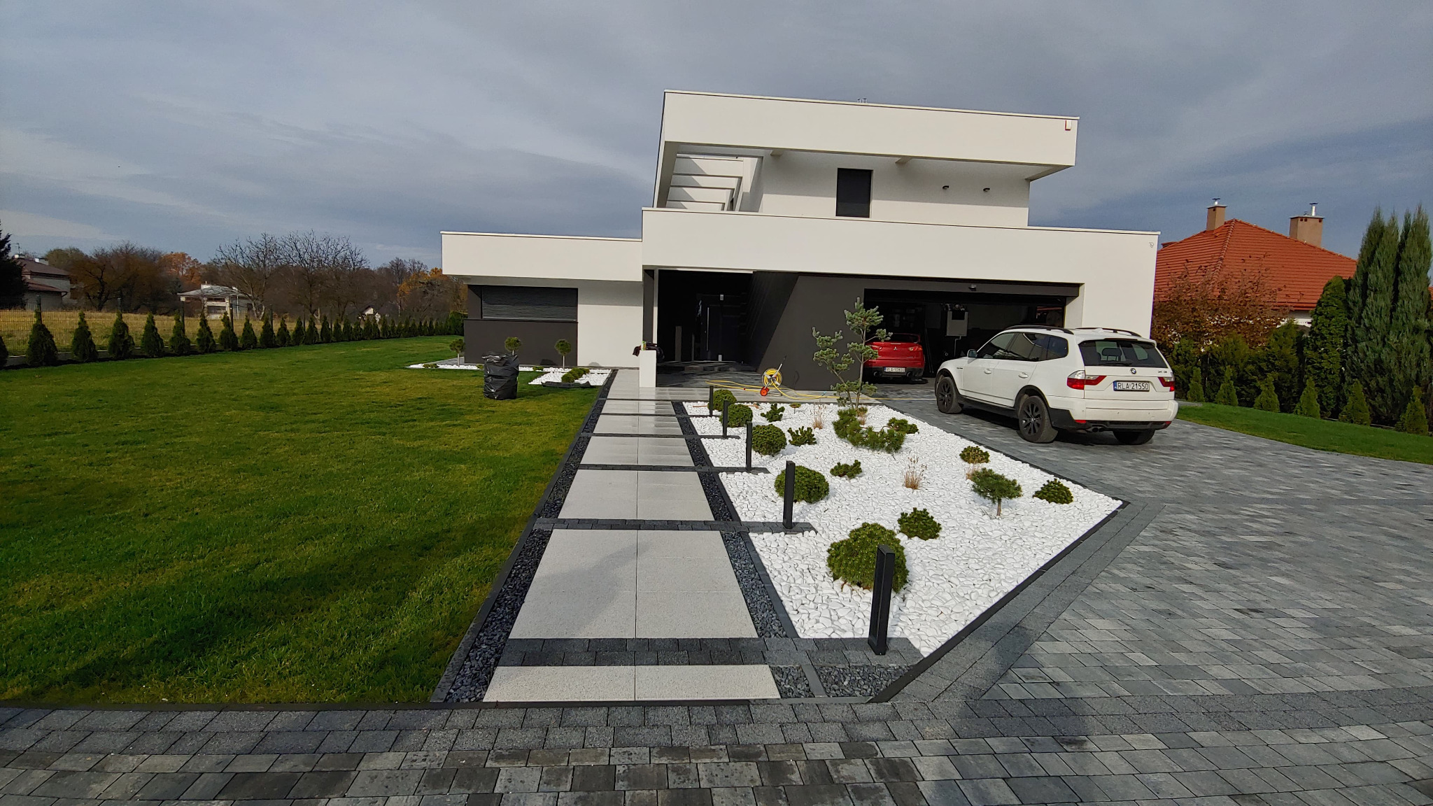 Nowoczesny dom z zadbanym ogrodem, ścieżką z płyt betonowych obramowanych czarnym grysem, trawnikiem i podjazdem z szarej kostki. Białe kamyki dekoracyjne z niskimi krzewami i drzewkami iglastymi.