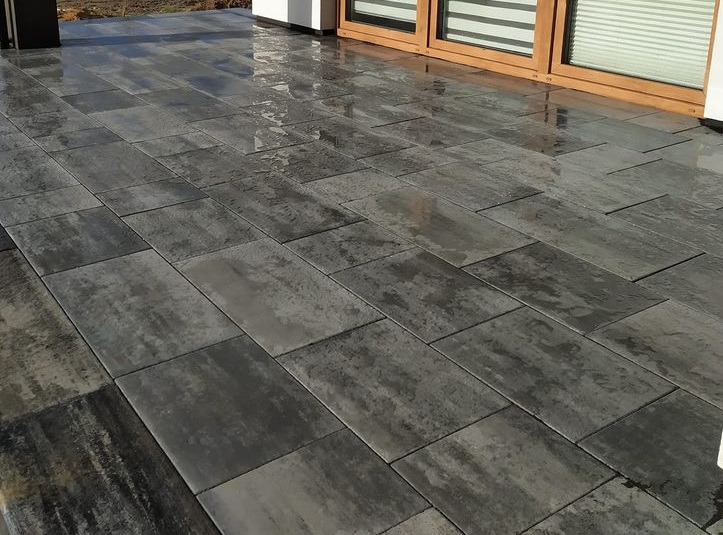 Ułożona, ciemnoszara nawierzchnia z prostokątnych płyt przy oknie z drewnianą ramą, lekko mokra od deszczu.