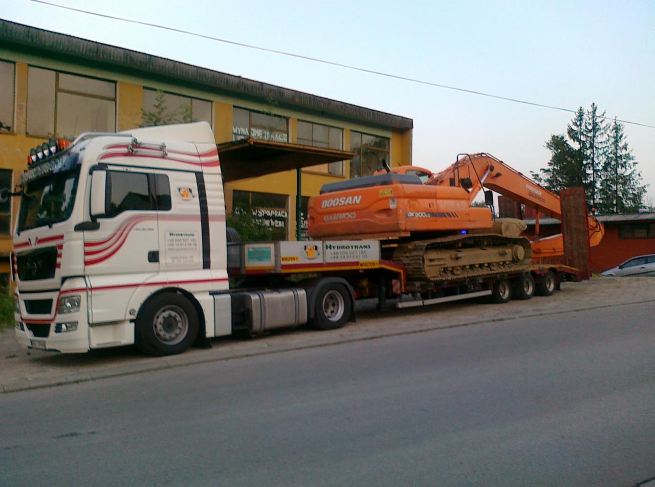 Biała ciężarówka z czerwonymi pasami przewozi pomarańczową koparkę Doosan na niskopodwoziowej platformie, widoczny budynek w tle.