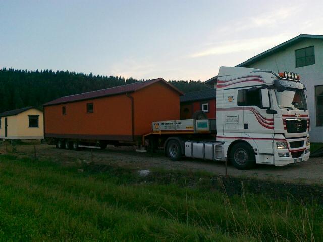 Ciężarówka niskopodwoziowa przewozi pomarańczowy, prefabrykowany budynek modułowy na platformie, z zielonymi drzewami w tle.