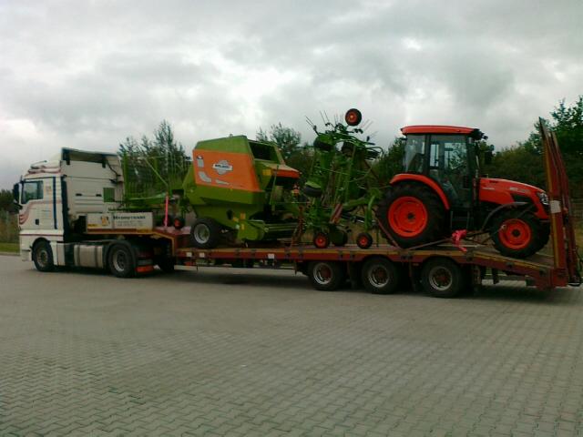 Ciężarówka z naczepą przewożąca pomarańczowy traktor, zieloną prasę rolniczą i przetrząsacz karuzelowy na tle pochmurnego nieba.